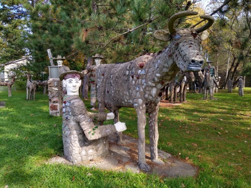 A stone sculpture of a person milking a cow, surrounded by other stone figures in a grassy area.