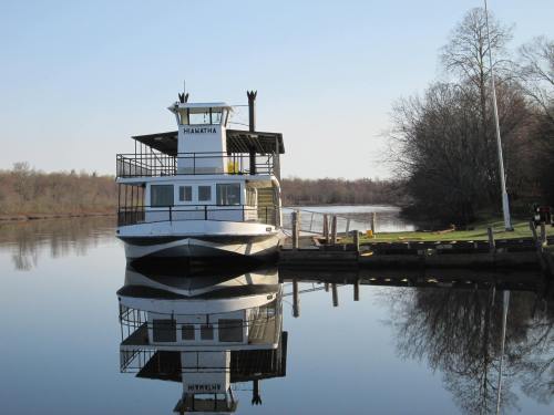 A boat docked by a calm river, reflecting on the water, surrounded by trees and a clear blue sky.