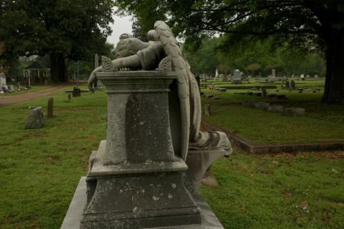 A weathered angel statue rests on a gravestone in a serene cemetery, surrounded by green grass and distant tombstones.