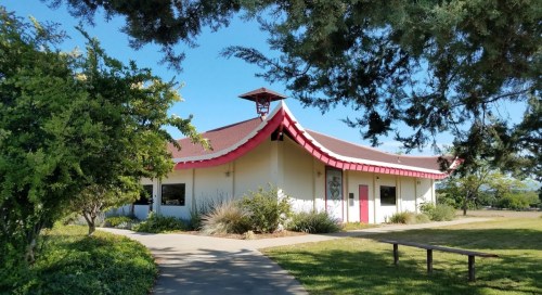 A modern building with a curved roof and red accents, surrounded by greenery and a clear blue sky.