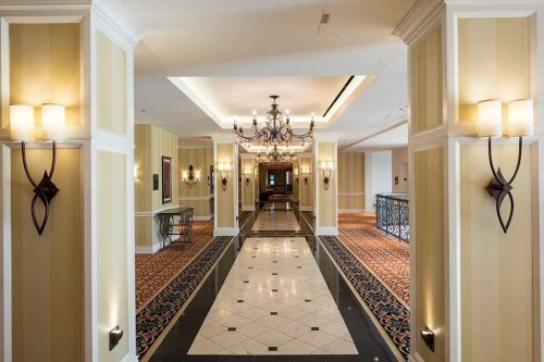 Elegant hotel hallway with patterned carpet, decorative lighting, and a chandelier, leading to a distant entrance.