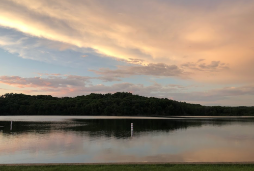 A serene lake at sunset, reflecting colorful clouds and surrounded by lush green trees.