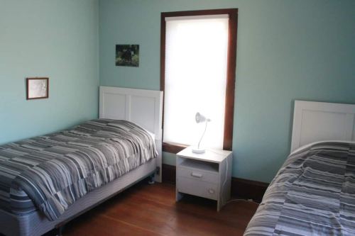 A light blue bedroom with two twin beds, a nightstand, and a lamp by a window.