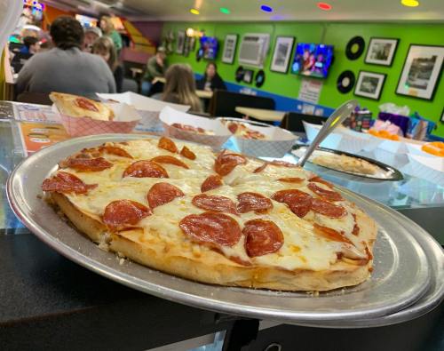 A close-up of a pepperoni pizza on a metal tray, with a colorful restaurant background and people dining.