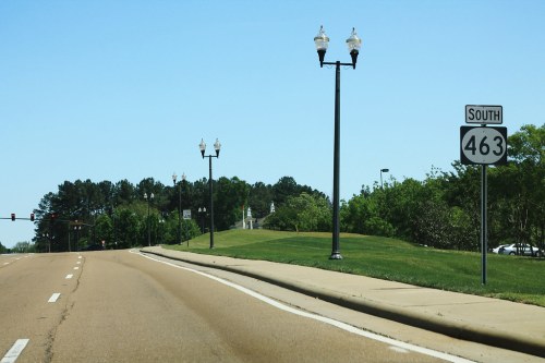 A clear road with a "South 463" sign, street lamps, and green trees lining the sides under a blue sky.