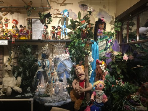 A colorful display of puppets and figures surrounded by greenery and decorations, creating a whimsical scene.