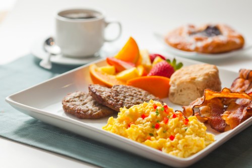 A breakfast plate with scrambled eggs, bacon, sausage, fruit, a biscuit, and a cup of coffee in the background.
