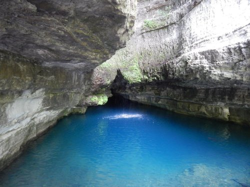 A serene cave with steep rock walls surrounding a vibrant blue pool of water. Sunlight filters in from above.