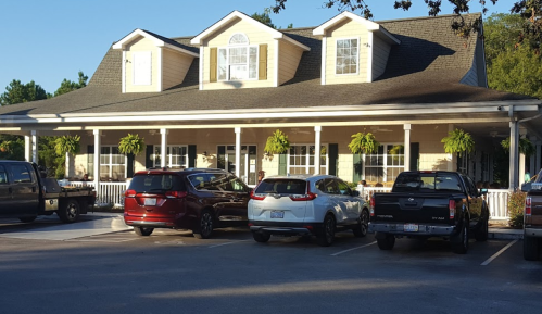 A two-story house with a porch, surrounded by parked cars and hanging plants, set in a sunny outdoor area.
