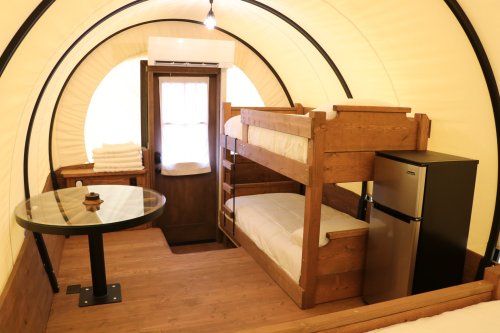 Interior of a cozy tent with bunk beds, a small table, and a fridge, featuring warm lighting and wooden accents.