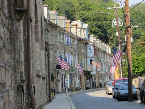 A charming street lined with stone buildings, featuring American flags and greenery in the background.