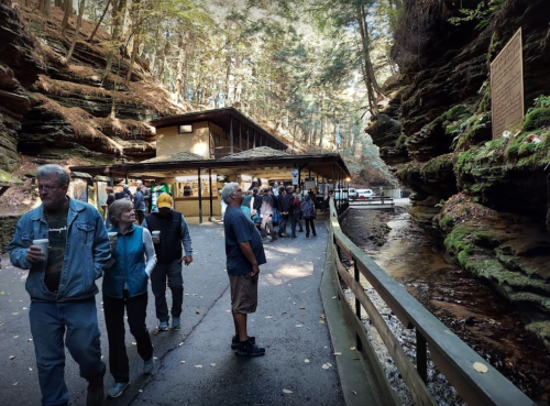 A group of people walking along a path near a building by a creek, surrounded by tall rock formations and trees.