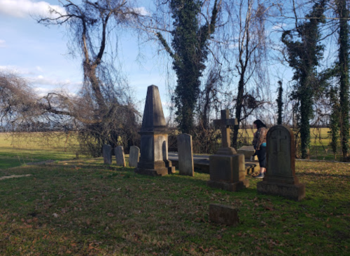 A person stands among old gravestones in a cemetery, surrounded by trees and a clear blue sky.