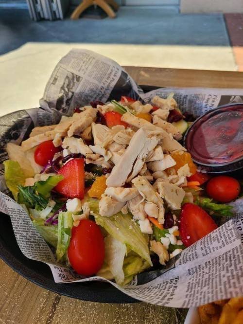 A colorful salad with lettuce, tomatoes, chicken, oranges, and cheese, served in a bowl lined with newspaper.