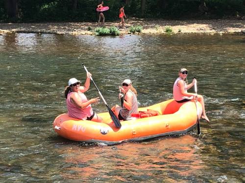 Three people in an orange raft paddle on a river, with a sandy bank and trees in the background.