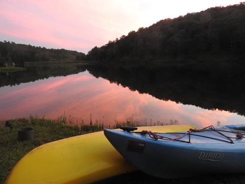 A serene lake at sunset, reflecting pink and orange hues, with two kayaks resting on the grassy shore.