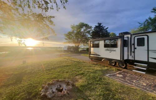 A scenic view of an RV parked near a lake at sunset, with a fire pit and trees in the foreground.