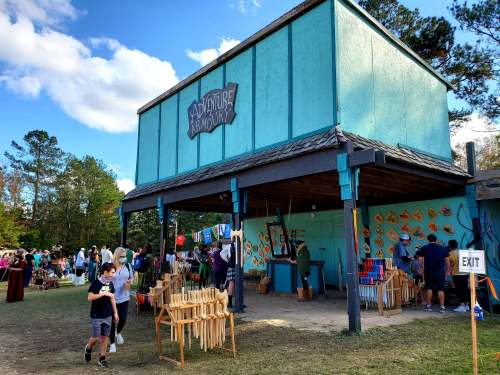 A colorful outdoor market scene with a blue building labeled "Adventure Armory" and people browsing various stalls.