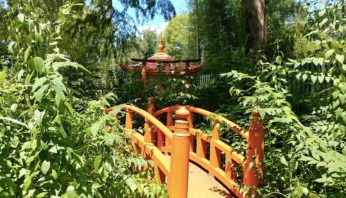 A vibrant orange bridge leads to a pagoda, surrounded by lush greenery and trees under a clear blue sky.