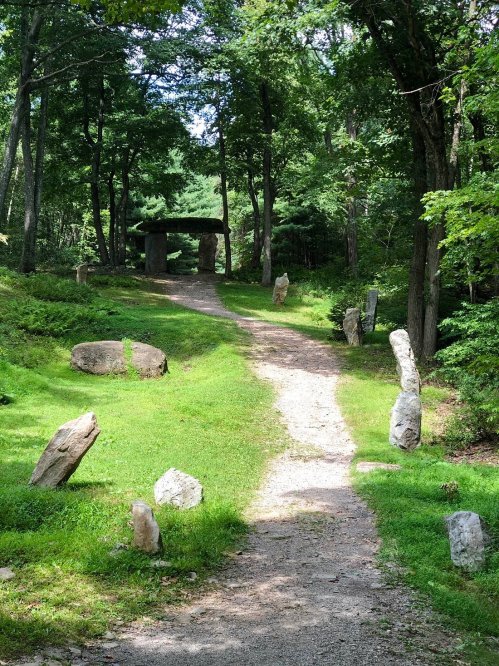 A serene pathway through a wooded area, flanked by stones, leading to a stone archway in the distance.