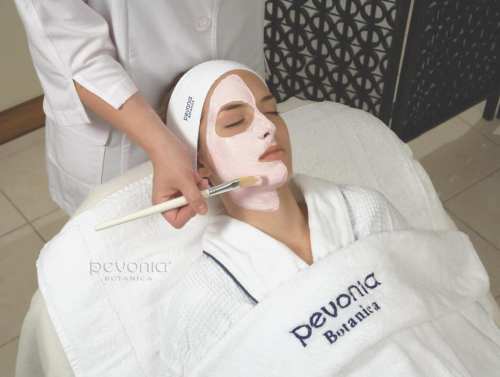 A woman receiving a facial treatment, with a therapist applying a mask using a brush. She is in a spa setting.