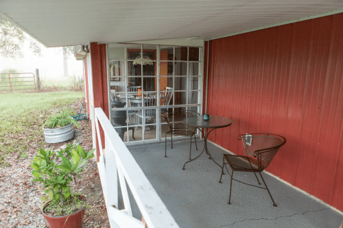 A cozy porch with a small table and chairs, overlooking a grassy area and a red wall.