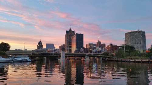 Sunset over a city skyline reflecting in the water, with buildings and boats visible along the shore.