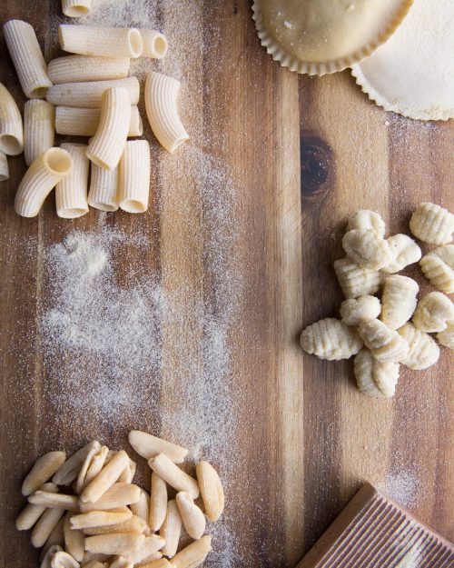 A wooden surface with various types of pasta: rigatoni, gnocchi, and ravioli, sprinkled with flour.