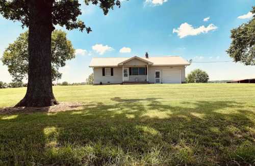 A single-story house with a garage, surrounded by green grass and trees under a blue sky with fluffy clouds.