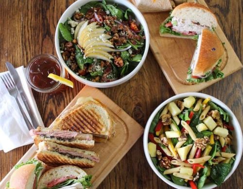 A variety of dishes on wooden platters, including salads, sandwiches, and a drink, arranged on a rustic table.