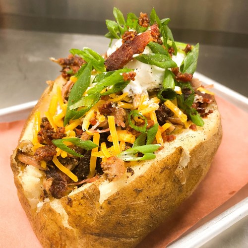 A loaded baked potato topped with cheese, bacon, green onions, and a dollop of sour cream on a metal tray.