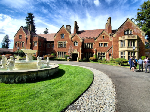 A grand brick mansion with a fountain in front, surrounded by lush green lawns and a clear blue sky.