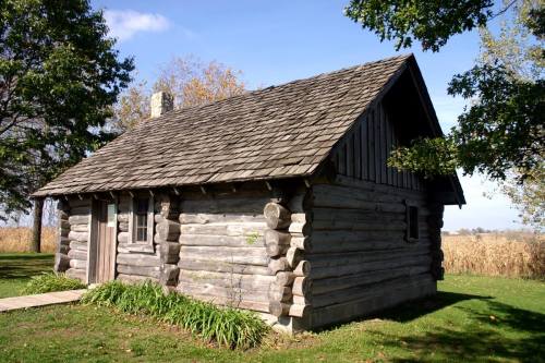 A rustic log cabin with a sloped roof, surrounded by greenery and trees under a clear blue sky.