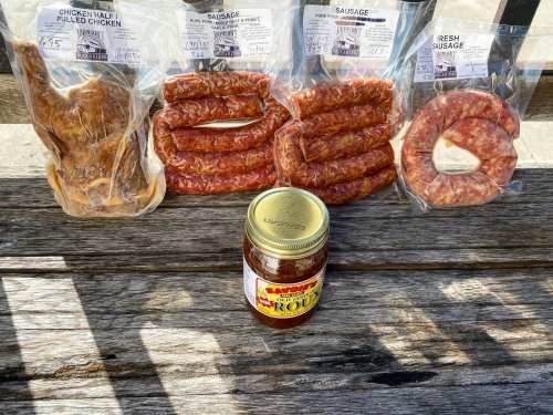 A wooden table displays packaged meats: pulled chicken, sausages, and a jar of barbecue sauce.
