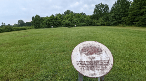 A grassy field with a weathered sign in the foreground, surrounded by trees in the background under a cloudy sky.