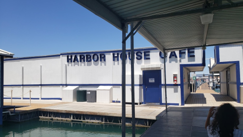 A waterfront view of the Harbor House Cafe with a clear blue sky and a dock in the foreground.