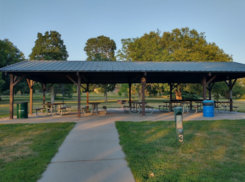 A covered picnic area with tables in a park, surrounded by grass and trees, with a path leading through.