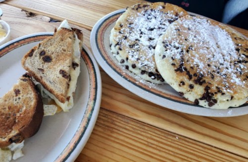A plate of fluffy pancakes with chocolate chips and powdered sugar, alongside a slice of toasted bread.