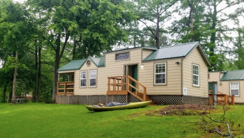 A cozy cabin with a green roof, surrounded by trees, and a kayak resting on the grass in front.