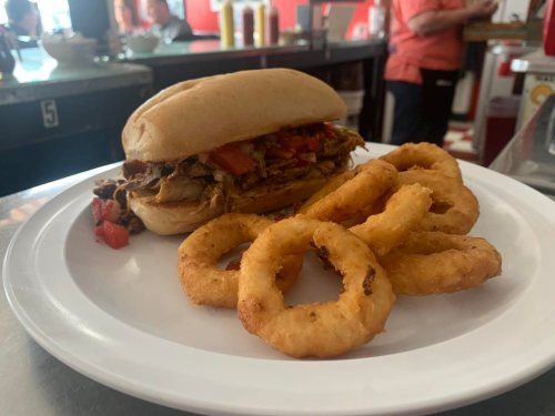 A plate with a sandwich filled with shredded meat and tomatoes, accompanied by crispy onion rings.