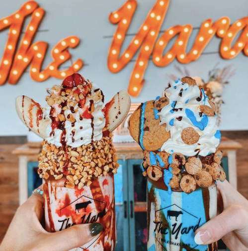 Two extravagant milkshakes topped with whipped cream, cookies, nuts, and a cherry, held in front of a lit sign reading "The Yard."