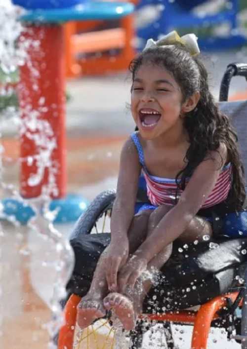 A joyful girl in a wheelchair splashes in water at a playground, smiling and enjoying the moment.