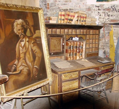 A vintage desk with a chair, surrounded by books, and a large portrait of an older man in a frame.