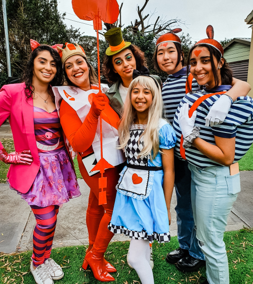 A group of six people in colorful costumes, including Alice and the Queen of Hearts, posing together outdoors.