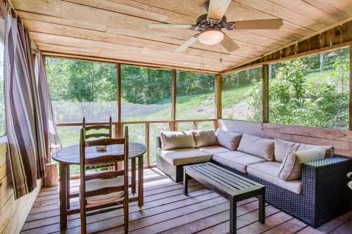 Cozy screened porch with a sofa, table, and chairs, surrounded by greenery and natural light.