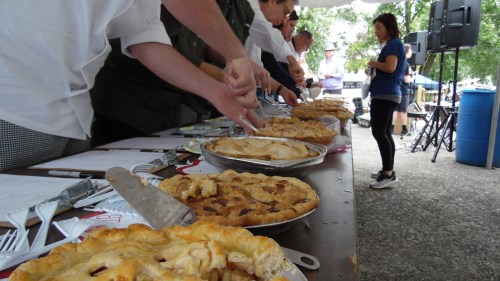 A pie judging event with several pies on a table and judges preparing to taste them.