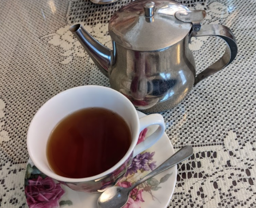 A silver teapot beside a floral teacup filled with tea on a lace tablecloth.
