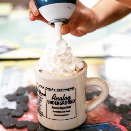 A hand holds a whipped cream dispenser over a mug labeled "Analog Underground," ready to top a drink.