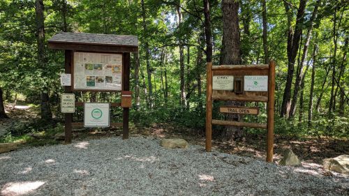 Trailhead sign in a wooded area, featuring maps and information about the trail, with a "Trail Open" sign.