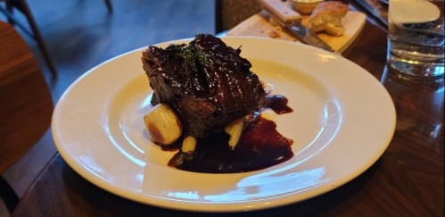 A plated dish featuring a slice of beef with a rich sauce, garnished with herbs, served on a white plate.